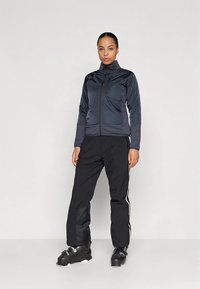 Granatowa kurtka na zamek z wysokim kołnierzem, w połączeniu z czarnymi spodniami z białym paskiem po boku. Model nosi czarne buty narciarskie.