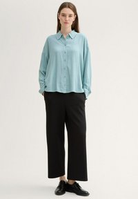 Camisa de botones de manga larga azul claro combinada con pantalones anchos negros y zapatos negros con cordones, de pie contra un fondo neutro.