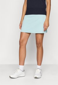 Světle modrá žebrovaná tenisová sukně s rozšířeným střihem, spárovaná s bílými sportovními botami a strukturovanými kotníkovými ponožkami.