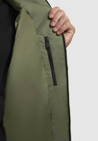Helles olivengrünes Jacket mit glatter Textur, das eine Reißverschlusstasche auf der inneren linken Seite und schwarze Reißverschlussbeschläge aufweist.