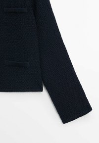 Manche et partie du corps d'un pull en maille texturée bleu marine foncé avec petite poche avant sur fond blanc.
