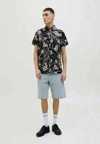 Jovem a usar uma camisa preta de manga curta com padrão floral, calções de ganga azul claro, meias brancas e sapatos pretos, de pé contra um fundo simples.