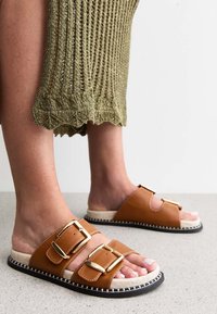 Sandalias de deslizamiento sintéticas marrones con dos grandes hebillas doradas, costuras blancas y una suela texturizada. Combinadas con una prenda larga de punto verde.