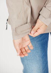 Beige jacka med handledsrem som har "W-PRO 10000" broderi. Slät textur, elastiskt mudd, hand placerad nära denimjeans.