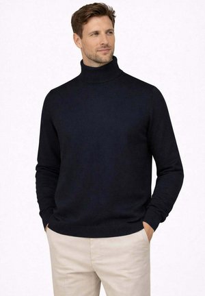 Uomo in piedi con le mani in tasca, indossando un maglione a collo alto nero e pantaloni beige su uno sfondo bianco.