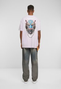 Lilla kortermet t-skjorte med et lionmotiv i sterke farger på baksiden, sammen med lysegrå vide jeans og hvite joggesko.