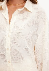 Camisa blanca con una textura deshilachada, que presenta un cuello y cierre de botones. Hecha de una tela ligera, con un patrón intrincado.