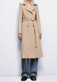 Gabardina beige de doble botonadura con cinturón, diseño entallado, hombreras y textura suave, combinada con vaqueros azules acampanados y zapatos negros.