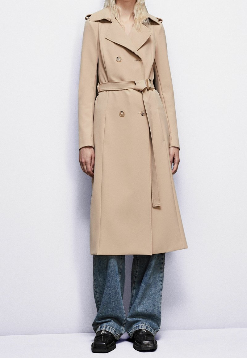 Gabardina beige de doble botonadura con cinturón, diseño entallado, hombreras y textura suave, combinada con vaqueros azules acampanados y zapatos negros.