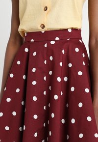 Maroonkjol med vita polka prickar, plisserad design och hög midja. Tillverkad av ett texturerat tyg som ger en strukturerad look.