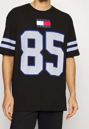 Czarny t-shirt Tommy Jeans z dużą białą i niebieską cyfrą 85 na przodzie, białe i niebieskie podwójne paski na rękawach, noszony przez stojącą osobę.