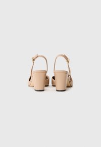  Béžové slingback blokové podpätky s remienkami na členku a zlatými prackami, zobrazené zozadu na jednoduchom bielom pozadí.