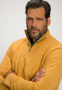 Gelber Reißverschluss-Pullover aus strukturiertem Stoff. Verfügt über einen kleinen Kragen und gerippte Bündchen. Das Muster weist subtile Farbvariationen auf.