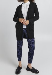 Musta avonainen neuletakki, ribattu pinta, kaksi etutaskua. Yhdistetty sinisiin leopardikuosisisiin housuihin ja mustiin loafereihin. Ylhäällä ei ole näkyviä kuvioita.