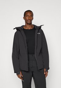 Schwarze gepolsterte Jacke mit Kapuze, ausgestattet mit Reißverschluss-Taschen und einem Logo-Akzent. Getragen über einem schwarzen Shirt und kombiniert mit schwarzen Hosen.