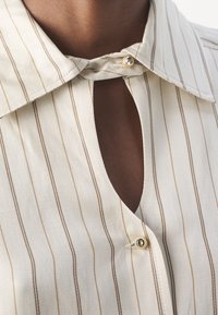 Camicia button-up di colore crema chiaro con strisce verticali marroni, dotata di un ampio colletto e un'apertura a chiave sul collo. Dettagli con bottoni dorati.