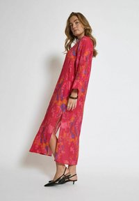Lang, pink kjole med farverige abstrakte mønstre, sideslids og lange ærmer. Letvægtsstof, ankel-længde, parret med sorte slingback hæle.