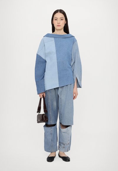 Denim outfit featuring a blue patchwork top with wide sleeves and loose-fitting pants. Holding a small, dark handbag.