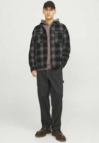 Een zwart-grijs geruite flanellen shirt over een bruine hoodie, gecombineerd met zwarte broek en bruine veterschoenen, tegen een effen achtergrond.
