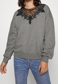 Sudadera gris con un detalle de encaje negro con patrones florales alrededor del escote, mangas largas y puños y dobladillo de canalé.