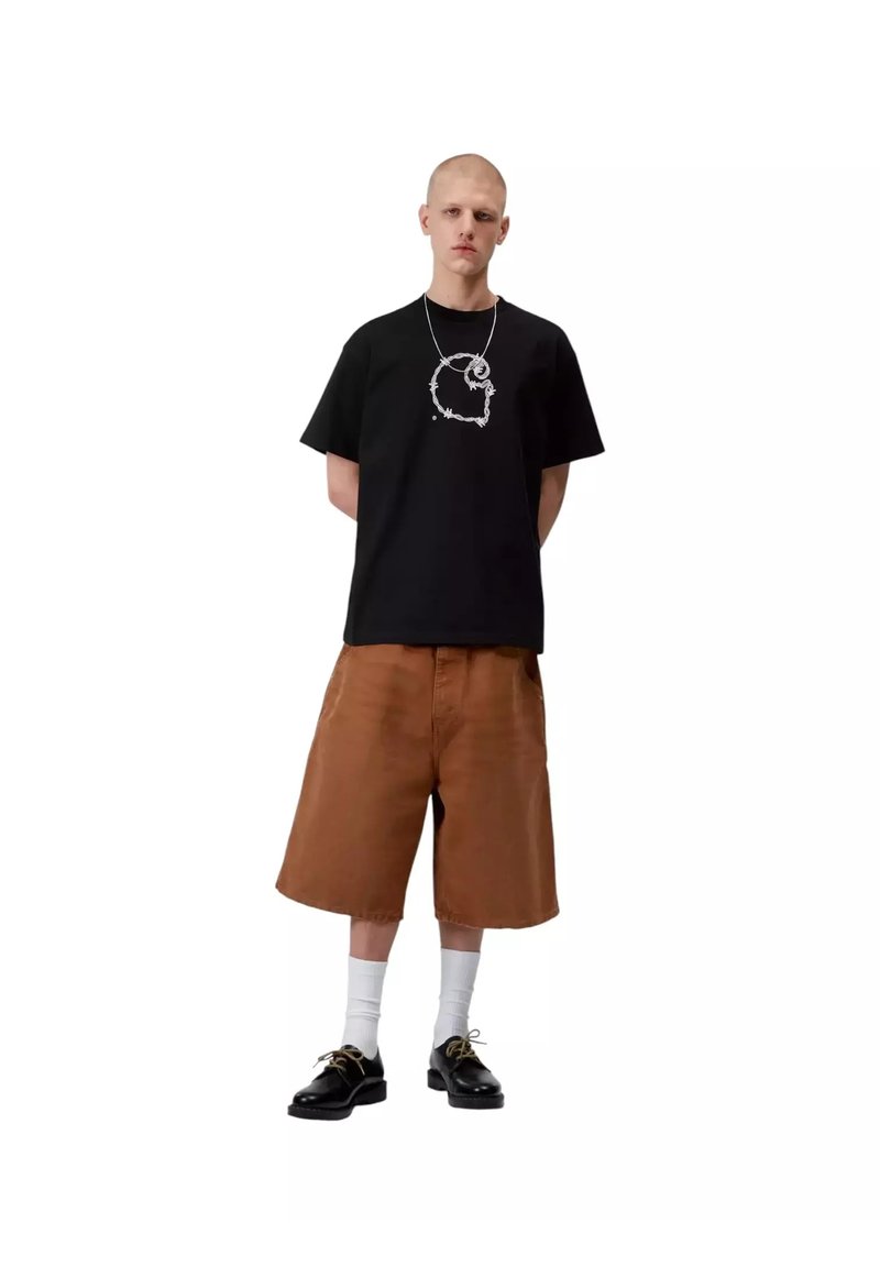 Junger Mann steht mit den Händen hinter dem Rücken, trägt ein schwarzes T-Shirt mit Grafik, braune weite Shorts, weiße Socken und schwarze Schuhe vor weißem Hintergrund.