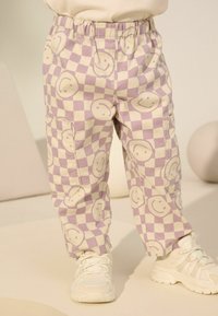 Lila- und cremefarbene karierte Cargo-Hosen mit Smiley-Gesicht-Mustern, elastischem Bund und seitlichen Taschen, kombiniert mit weißen Sneakern.