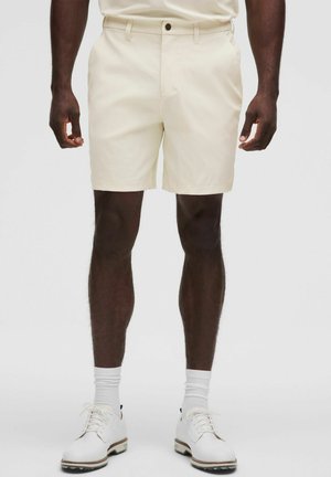 Beige shorts i en lige pasform med forlommer, knaplukning og bæltestropper, kombineret med hvide sko og sokker.