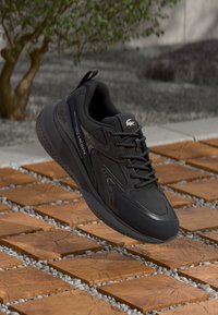 Black athletic sneakers with a smooth leather upper, textured accents, lace-up design, and a rubber sole, featuring the Lacoste logo.