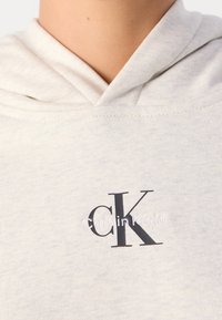 Gros plan d'un sweat à capuche beige clair avec le logo texte noir "cK" et blanc "Calvin Klein" sur la poitrine sous l'encolure.