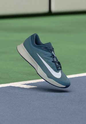 Blå Nike løbesko med hvid swoosh, der svæver over hjørnet af en tennisbane med grønne og blå overflader.