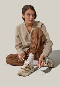 Beige gestrickte Strickjacke über einem hellbraunen Hemd, kombiniert mit braunen, schmal zulaufenden Hosen. Schuhwerk: Klobige Sneakers mit Stoff- und Wildlederakzenten, weiße Sohle.