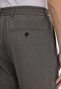 Primo piano di pantaloni grigi texture con vita elastica e un singolo bottone sulla tasca posteriore, indossati da una persona in camicia bianca.