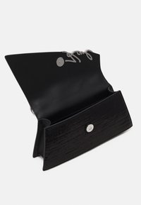 Pochette rectangulaire noire avec devant texturé, rabat ouvert montrant une fermeture magnétique et un détail signature argenté à l’intérieur du rabat.