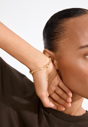 Detail osoby s uhladenými vlasmi učesanými dozadu, nosiaci zlatý retiazkový náramok, odpočívajúci rukou za uchom, oblečený v tmavej košeli.