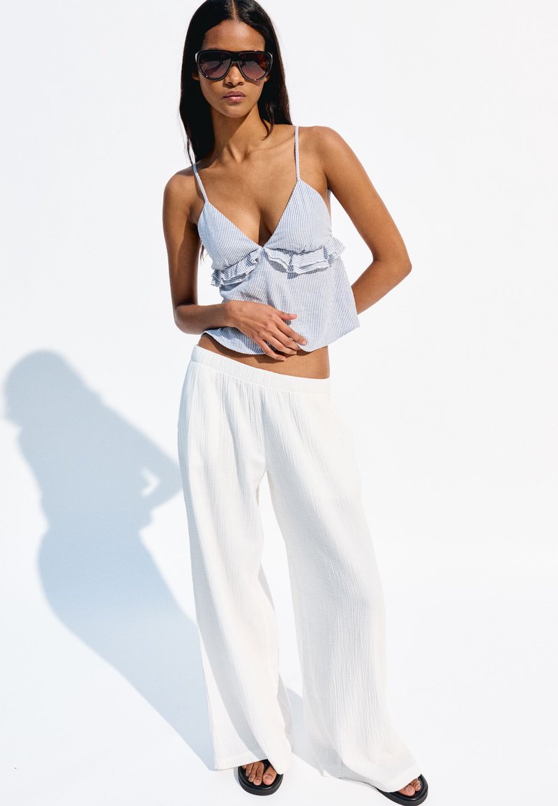 Pruhovaný crop top s volánkovým detailem v modré a bílé barvě, kombinovaný s bílými širokými kalhotami. Sluneční brýle a černé sandály doplňují vzhled.