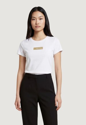 Giovane donna con i capelli lunghi e neri indossa una maglietta bianca a maniche corte con un logo rettangolare beige e pantaloni neri, in piedi davanti a uno sfondo grigio.