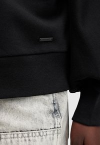 Sudadera negra con tejido texturizado, dobladillo acanalado y una pequeña etiqueta metálica con 'ALFAMATE' en la parte frontal, combinada con jeans de mezclilla claros.
