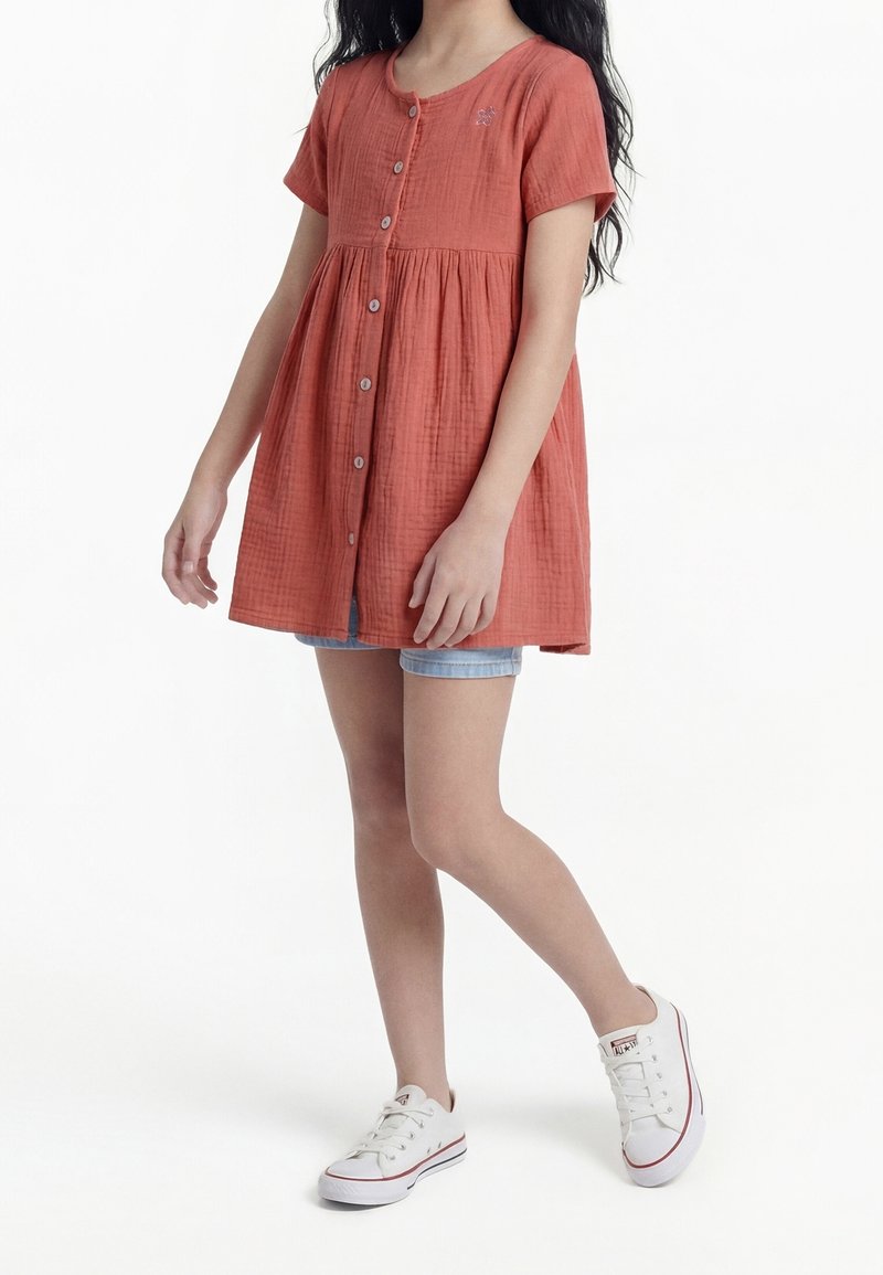 Fille portant une robe courte à manches courtes boutonnée couleur rouille par-dessus un short en jean et des baskets blanches basses, debout avec une jambe croisée.