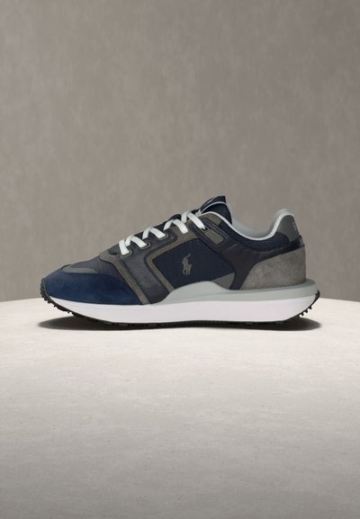 Σκούρα μπλε, γκρι και μαύρο αθλητικό παπούτσι με λευκά κορδόνια, μαλακή σόλα και μικρό κεντημένο λογότυπο στο πλάι, εμφανίζεται σε ουδέτερο φόντο.