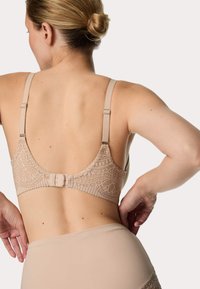 Béžová krajková podprsenka s nastavitelnými ramínky, květinovým vzorem na zadní straně a zapínáním na háček. Hladká textura, minimalistický design.