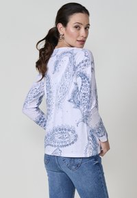 Mujer con cabello castaño en una coleta, vistiendo una camisa blanca de manga larga con un patrón de paisley azul y jeans azules, de espaldas con la cabeza girada.