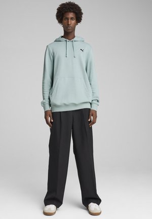 Sweatshirt vert clair avec capuche et poche avant, arborant un petit logo noir. Associez-le avec un pantalon noir ample et des baskets blanches.