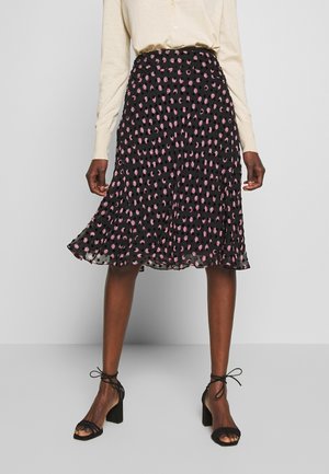 Femme portant une jupe noire jusqu'aux genoux avec des pois roses, un haut beige à manches longues et des sandales noires à talons avec des brides à la cheville.