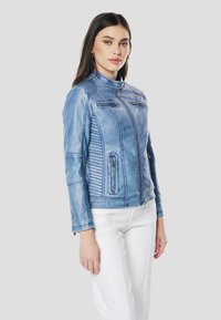 Blauwe jas van nepleer met een opstaande kraag, ritssluiting aan de voorkant en twee borstzakken met rits. Heeft verticale naaddetails en geribbelde accenten.