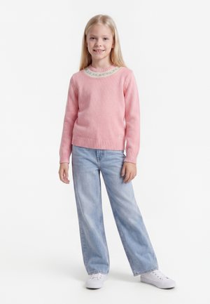 Jeune fille aux longs cheveux blonds portant un pull rose, un jean bleu clair et des baskets blanches, debout devant un fond blanc.