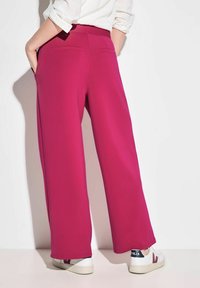 Fuchsia wijde broek met een gladde textuur, elastische tailleband en twee achterzakken, gecombineerd met witte sneakers, gezien van achteren.