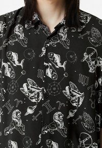 Camisa negra abotonada con ilustraciones en blanco de temática astrológica y zodiacal, que presenta leones, peces, manos y símbolos celestiales en un patrón repetido.