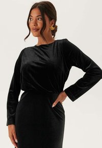 Robe en velours noir à manches longues et col rond. Texture lisse, silhouette ajustée et drapés subtils au niveau de la taille.