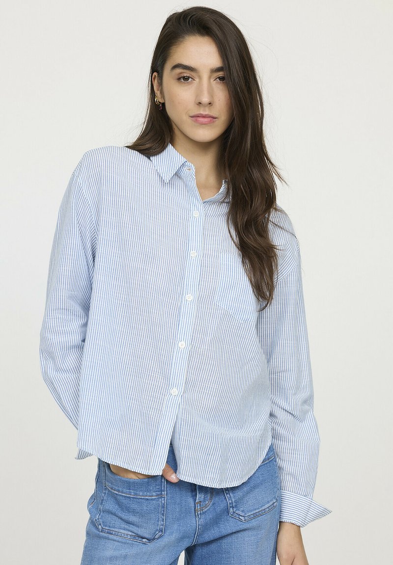 Chemise boutonnée rayée bleu clair avec une coupe décontractée, manches longues et une poche poitrine, associée à un jean bleu. Texture de tissu douce.