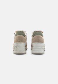 Baskets de sport beiges et grises avec des textures en daim et en cuir, présentant un accent vert et une marque en relief sur le talon. Semelle blanche épaisse.